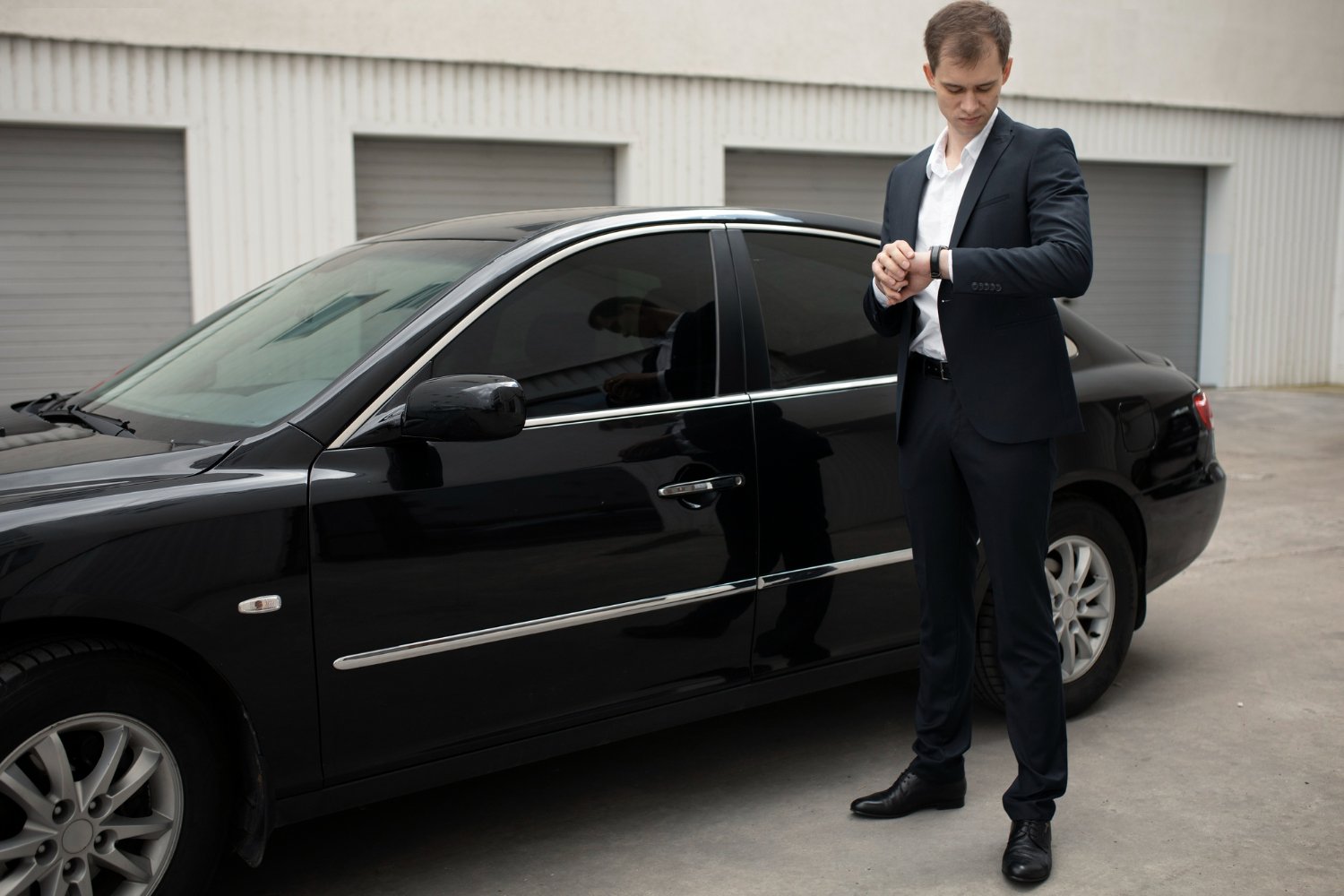 Professional chauffeur in suit checking a wristwatch beside a black sedan — hourly service is built around time